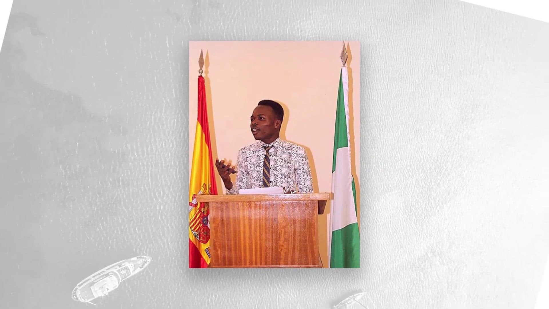 Fatola speaking at a lectern between the flags of Spain and Nigeria